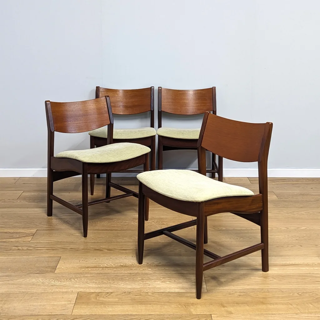 This fine set of four teak dining chairs have such a distinctive design. It's taken quite the investigation to attribute them. We initially thought they were British in origin but we're now confident they're Danish, made by Glostrup Modelfabrik and date from the 1960s. They're top quality with a heavy, solid construction but despite their stocky, wide stance, there are some superb details. The wave of the side bars and the contours of the wooden back are beautiful. They have been reupholstered in recent years in a neutral, pale textured fabric that has a hint of green to it. The light fabric actually complements the rich, warm teak well and it's in fantastic order but it would be an easy inexpensive job to recover them if it didn't suit. It would be worthwhile as they're a super set with provenance and heritage.  