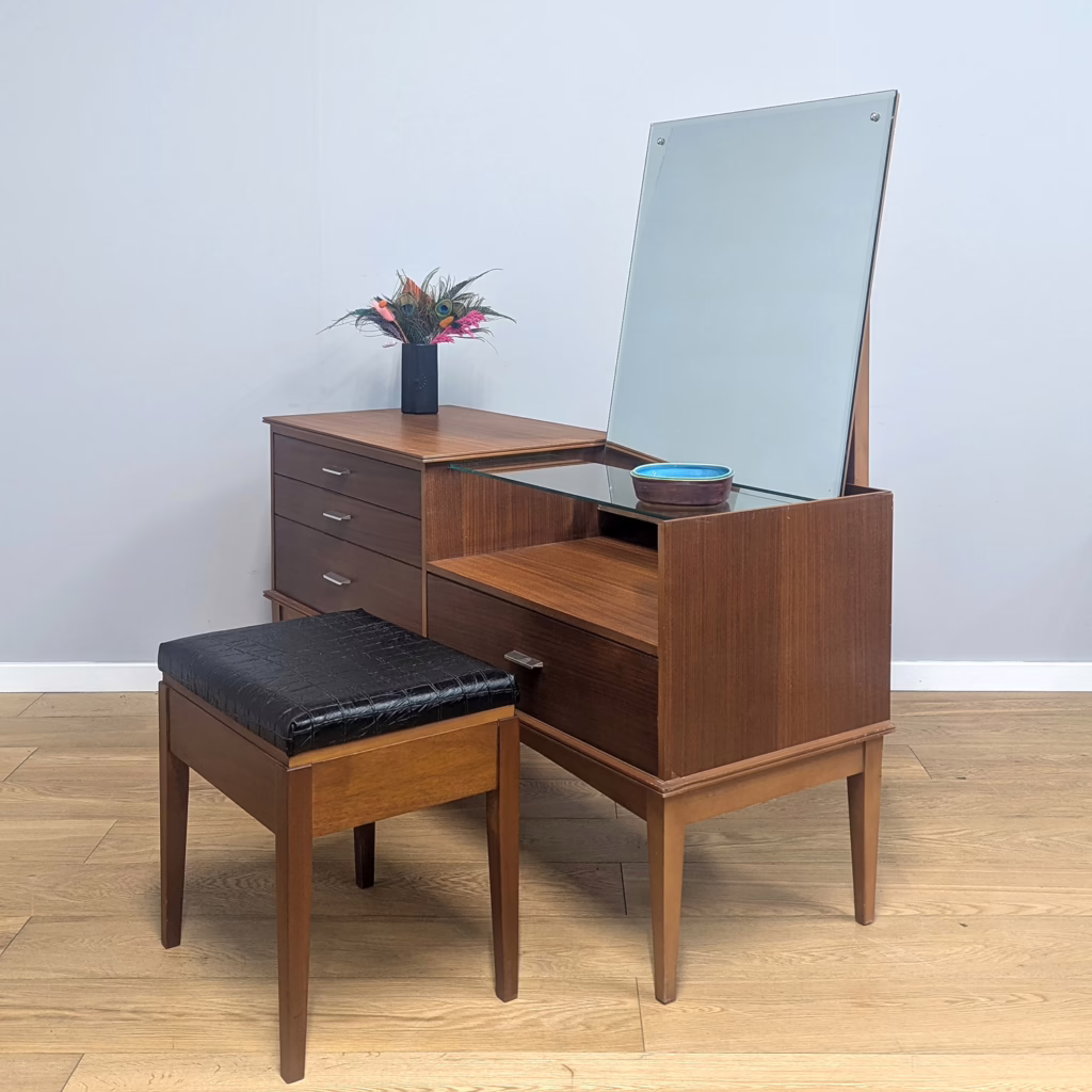 This fine, midcentury dressing table / vanity stand has had us confused. The design is so distinctive and the build of a superior quality, it must have been made by a high-end, reputable maker. But we can't find another. Our educated hunch is it's a rarely seen Uniflex piece, perhaps a short production run. The metal pull handles, the detail of the lower trim and the two tones of wood lead us to Uniflex. Indeed, looking back at a tallboy we previously stocked, there are definite similarities. Part chest of drawers, part dressing table, it was made for the bedroom / dressing room and creates a dedicated vanity spot for getting ready. We love the generous angled mirror and the recessed area, partially covered with a glass shelf for perfumes and potions. Although asymmetrical, it's perfectly balanced. A cracking find!